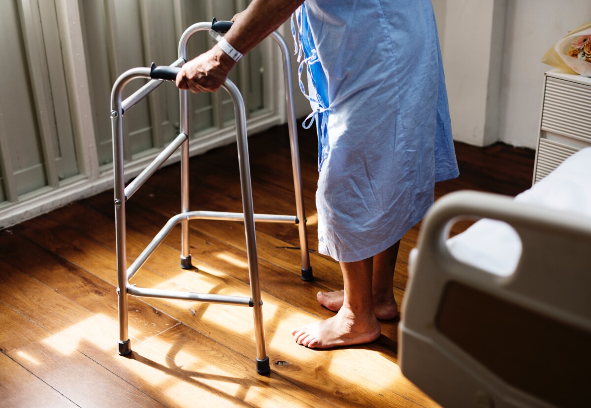 Personne âgée se déplaçant à son domicile avec un déambulateur, près d'un lit médicalisé